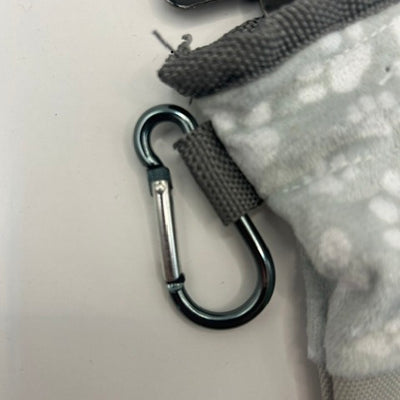 A close-up of a silver metal carabiner clipped onto a gray fabric strap attached to a light gray patterned fleece pouch.