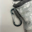 A close-up of a silver metal carabiner clipped onto a gray fabric strap attached to a light gray patterned fleece pouch.